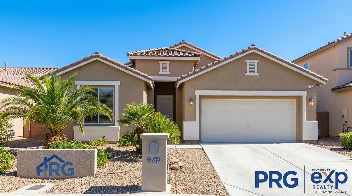 A tan stucco house with a tiled roof, white garage door, palm trees, and a concrete driveway.